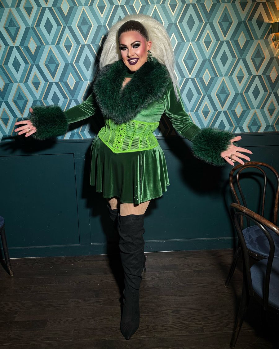 Abby Cummings in green velvet and faux fur cuffs, arms open wide in a Boston bar